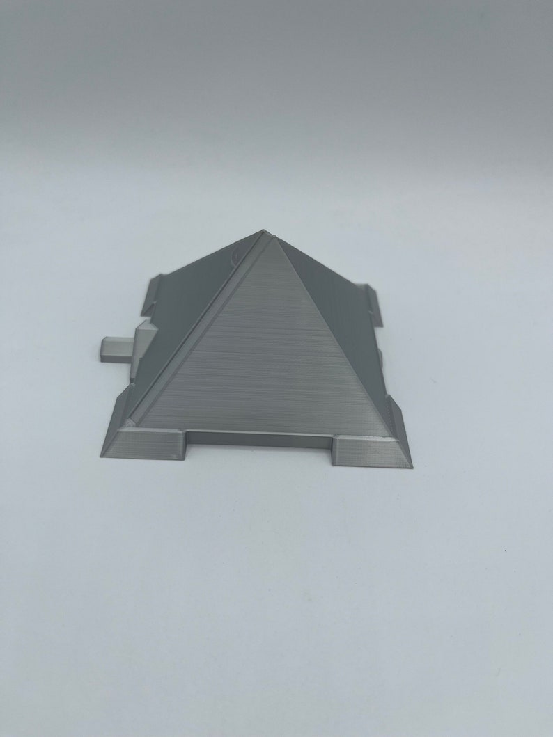 Puede incluir: Modelo de pir&aacute;mide gris impreso en 3D con una superficie texturizada. La pir&aacute;mide tiene una base rectangular con peque&ntilde;as extensiones rectangulares a cada lado. La parte superior de la pir&aacute;mide es un &aacute;pice puntiagudo.