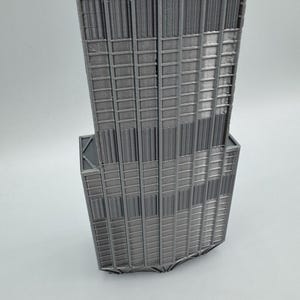 May include: A 3D printed model of a skyscraper featuring a grey finish. The structure displays a vertical grid pattern with recessed windows and a stepped architectural design, showcasing the layered texture typical of additive manufacturing.