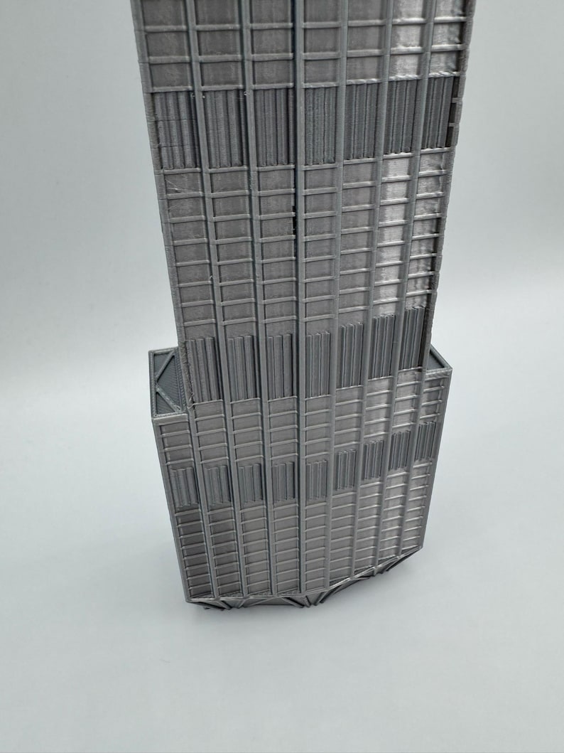 May include: A 3D printed model of a skyscraper featuring a grey finish. The structure displays a vertical grid pattern with recessed windows and a stepped architectural design, showcasing the layered texture typical of additive manufacturing.