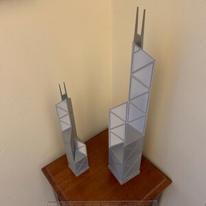 May include: Two gray architectural models of skyscrapers, one taller than the other, are displayed on a dark wooden table. The models have a geometric design with triangular and rectangular shapes. A ruler is placed in front of the models.