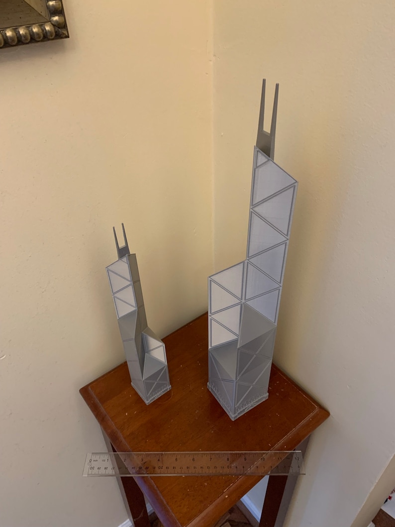 May include: Two gray architectural models of skyscrapers, one taller than the other, are displayed on a dark wooden table. The models have a geometric design with triangular and rectangular shapes. A ruler is placed in front of the models.