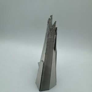 May include: A 3D printed model of a skyscraper, made of a silver-colored plastic. The model is detailed and has a sharp, angular design.