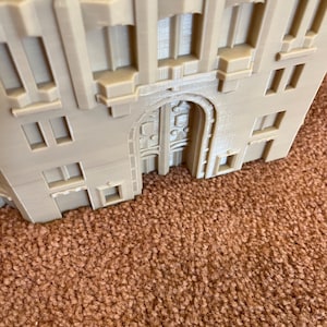 May include: A beige 3D printed model of a building with an arched doorway and multiple windows. The building has a detailed facade with decorative elements.