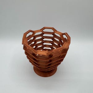 May include: A brown, open-weave decorative bowl with a honeycomb design. The bowl has a wide, open top and a pedestal base. The geometric pattern creates a modern, artistic look. The bowl is made of a solid material.