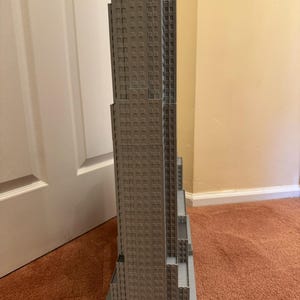 May include: A gray scale model of the Empire State Building. The model is a detailed replica of the iconic skyscraper, with numerous windows and architectural details. The model is placed on a gray base and sits on a brown carpet.