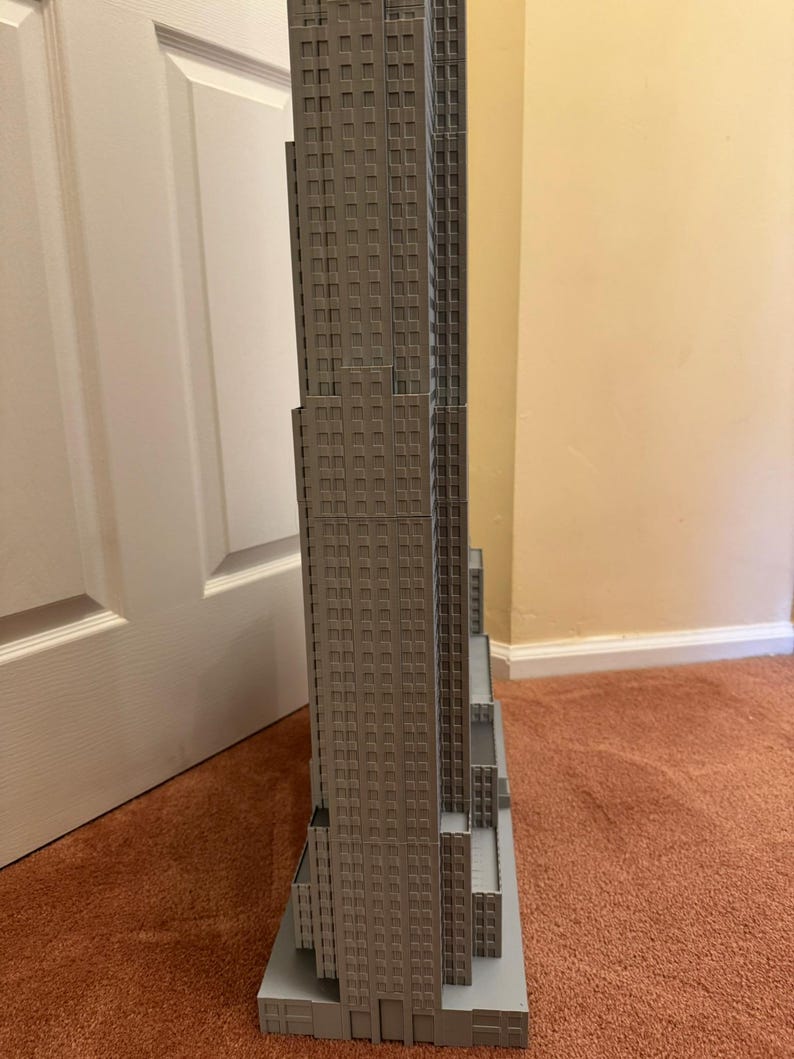 May include: A gray scale model of the Empire State Building. The model is a detailed replica of the iconic skyscraper, with numerous windows and architectural details. The model is placed on a gray base and sits on a brown carpet.
