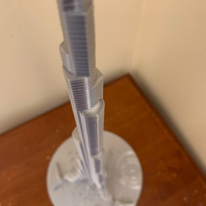 May include: A white 3D printed model of a skyscraper on a white base.