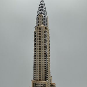 May include: A detailed model of the Chrysler Building, a prominent Art Deco skyscraper. The building features a beige facade with dark window details and a silver, tiered crown. The model is set against a plain background.