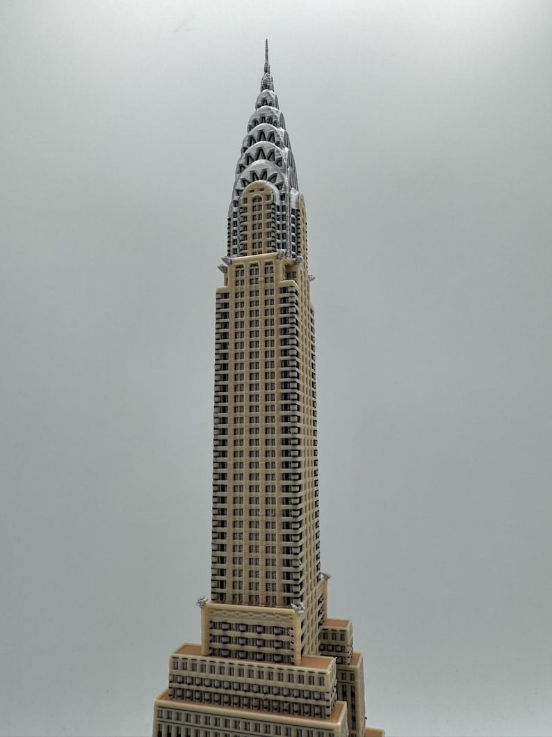May include: A detailed model of the Chrysler Building, a prominent Art Deco skyscraper. The building features a beige facade with dark window details and a silver, tiered crown. The model is set against a plain background.