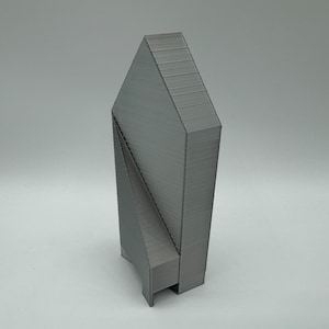 May include: A gray, 3D printed model of a building with a unique, angular design. The building has a tall, narrow shape with a pointed roof and a series of angled panels that create a geometric pattern.