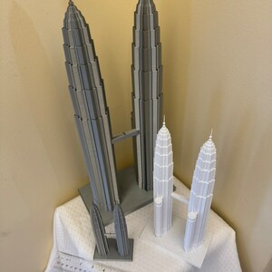 May include: Two sets of gray and white 3D printed models of the Petronas Towers. The larger set is gray and the smaller set is white. Both sets are on a white tablecloth with a ruler in the foreground.