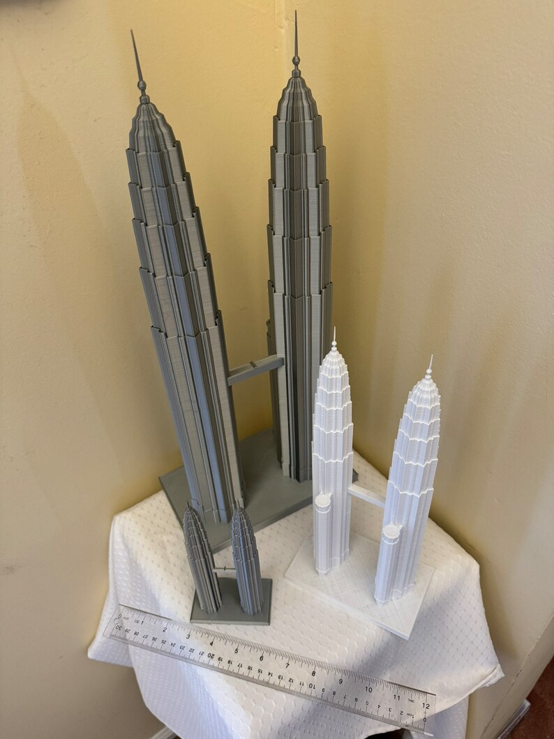 May include: Two sets of gray and white 3D printed models of the Petronas Towers. The larger set is gray and the smaller set is white. Both sets are on a white tablecloth with a ruler in the foreground.