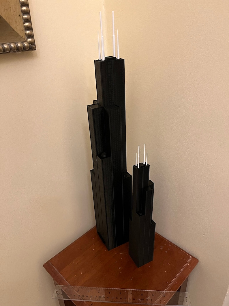 May include: Two black 3D printed models of skyscrapers with white antenna details. The models are on a wooden surface.