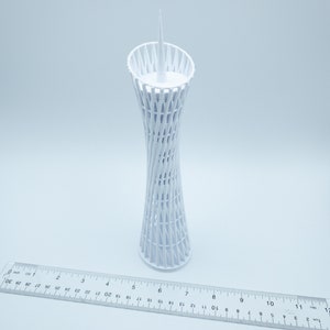 May include: White 3D printed model of a tall, slender tower with a lattice design. The tower is about 7 inches tall and has a pointed top.