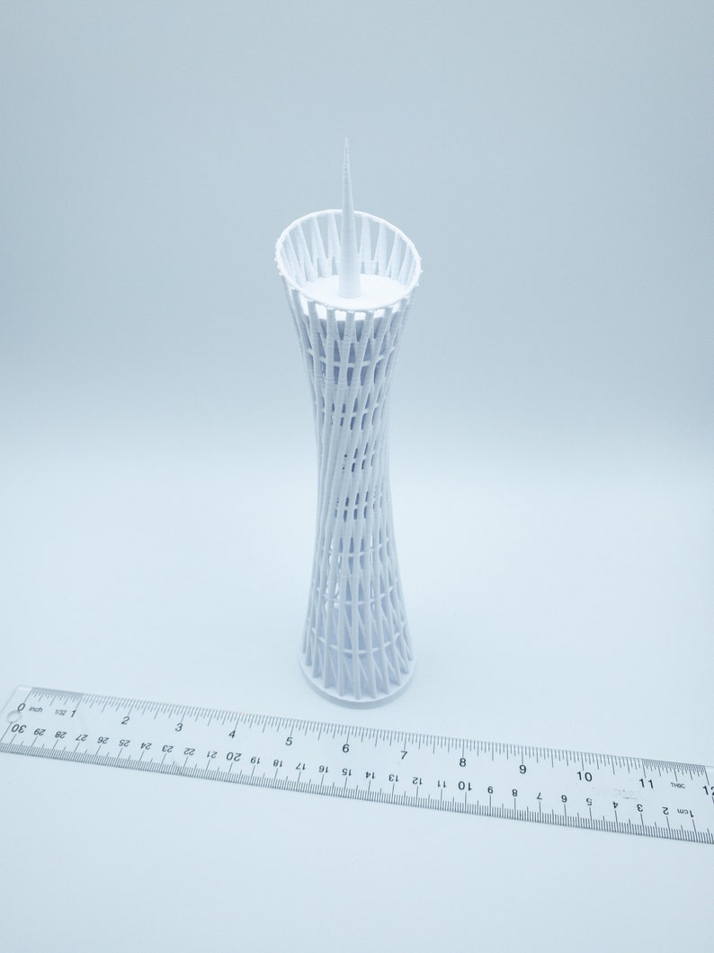 May include: White 3D printed model of a tall, slender tower with a lattice design. The tower is about 7 inches tall and has a pointed top.