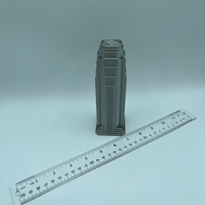 May include: A gray 3D printed model of a skyscraper with a detailed design. The model is about 3 inches tall and is sitting on a white surface next to a ruler.
