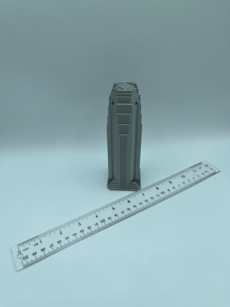 May include: A gray 3D printed model of a skyscraper with a detailed design. The model is about 3 inches tall and is sitting on a white surface next to a ruler.