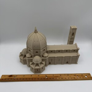 May include: A beige 3D-printed model of the Florence Cathedral, including the dome, side buildings, and bell tower. A wooden ruler is placed in front of the model, showing inch measurements. The model is detailed and sits on a white surface.