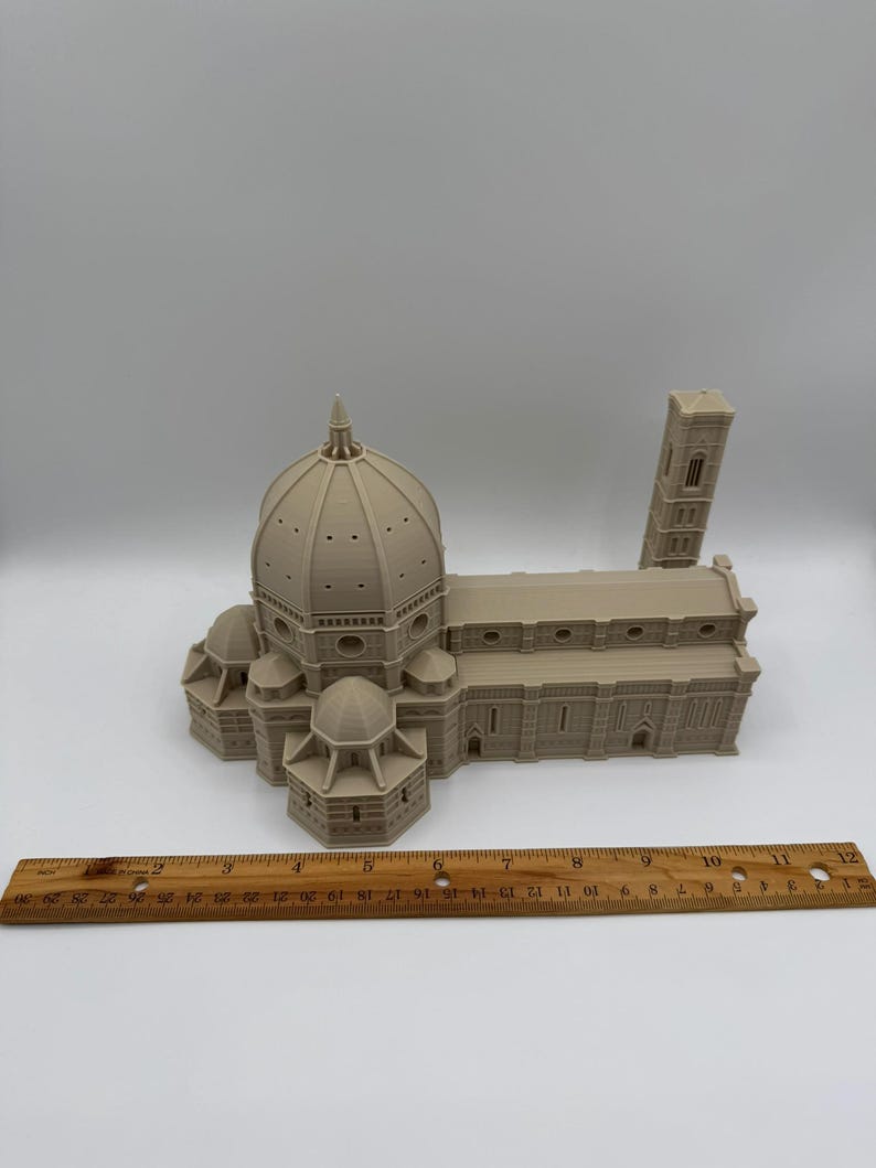 May include: A beige 3D-printed model of the Florence Cathedral, including the dome, side buildings, and bell tower. A wooden ruler is placed in front of the model, showing inch measurements. The model is detailed and sits on a white surface.