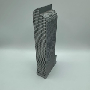 May include: A gray architectural model of a skyscraper. The building has a rectangular base and a tall, textured facade with a curved top. The model is made of a solid, matte gray material.