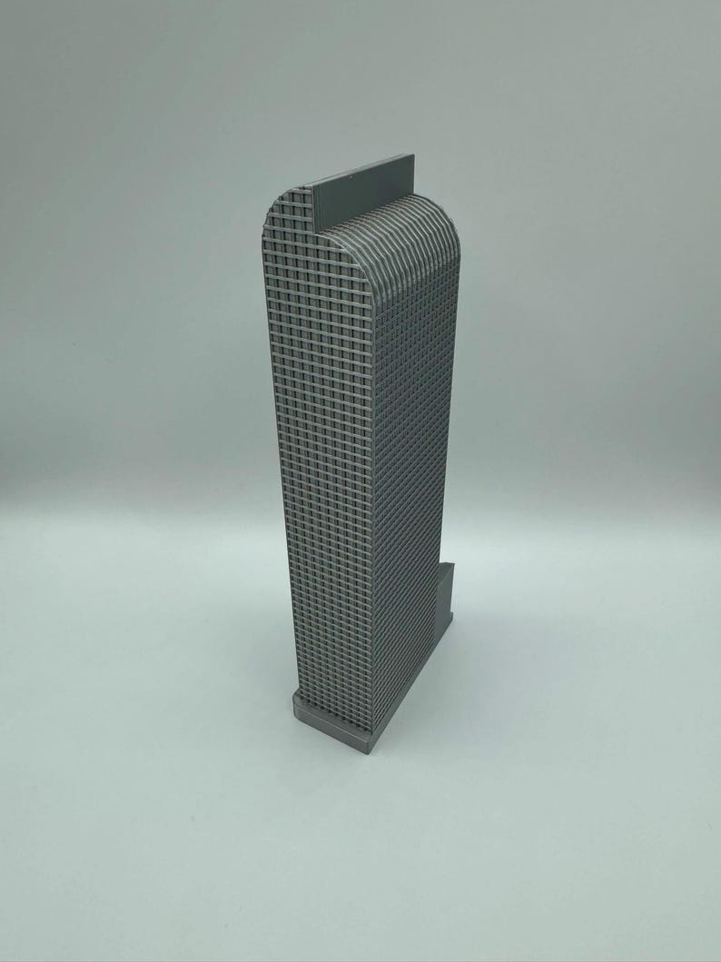 May include: A gray architectural model of a skyscraper. The building has a rectangular base and a tall, textured facade with a curved top. The model is made of a solid, matte gray material.