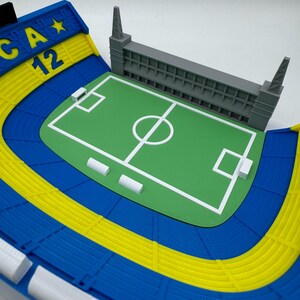May include: A detailed miniature stadium model with a green pitch, white markings, and grey seating. The stadium has blue and yellow seating areas, with the text "CA 12" on a blue and yellow structure.