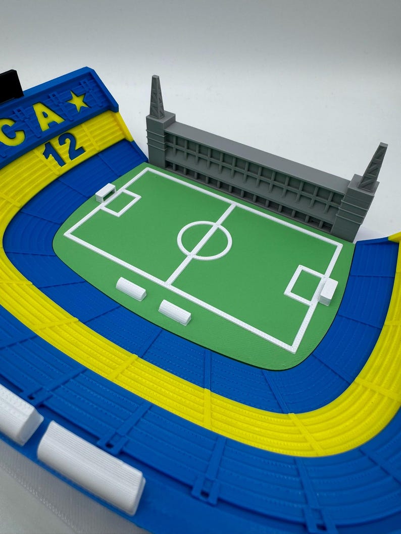 May include: A detailed miniature stadium model with a green pitch, white markings, and grey seating. The stadium has blue and yellow seating areas, with the text "CA 12" on a blue and yellow structure.