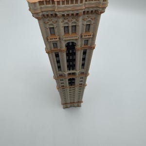 May include: A detailed miniature model of a tall building with a beige facade and brown accents. The building features multiple windows, decorative elements, and a detailed roof. The model is set against a plain white background.