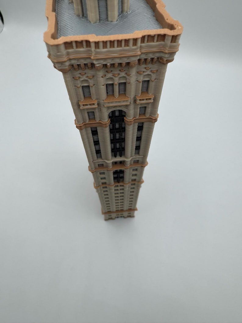 May include: A detailed miniature model of a tall building with a beige facade and brown accents. The building features multiple windows, decorative elements, and a detailed roof. The model is set against a plain white background.