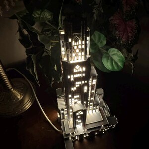 May include: A tall, dark, multi-tiered architectural lamp shaped like a skyscraper with numerous illuminated rectangular windows. The structure features intricate vertical detailing, a central tower, and a white power cord extending from the base.