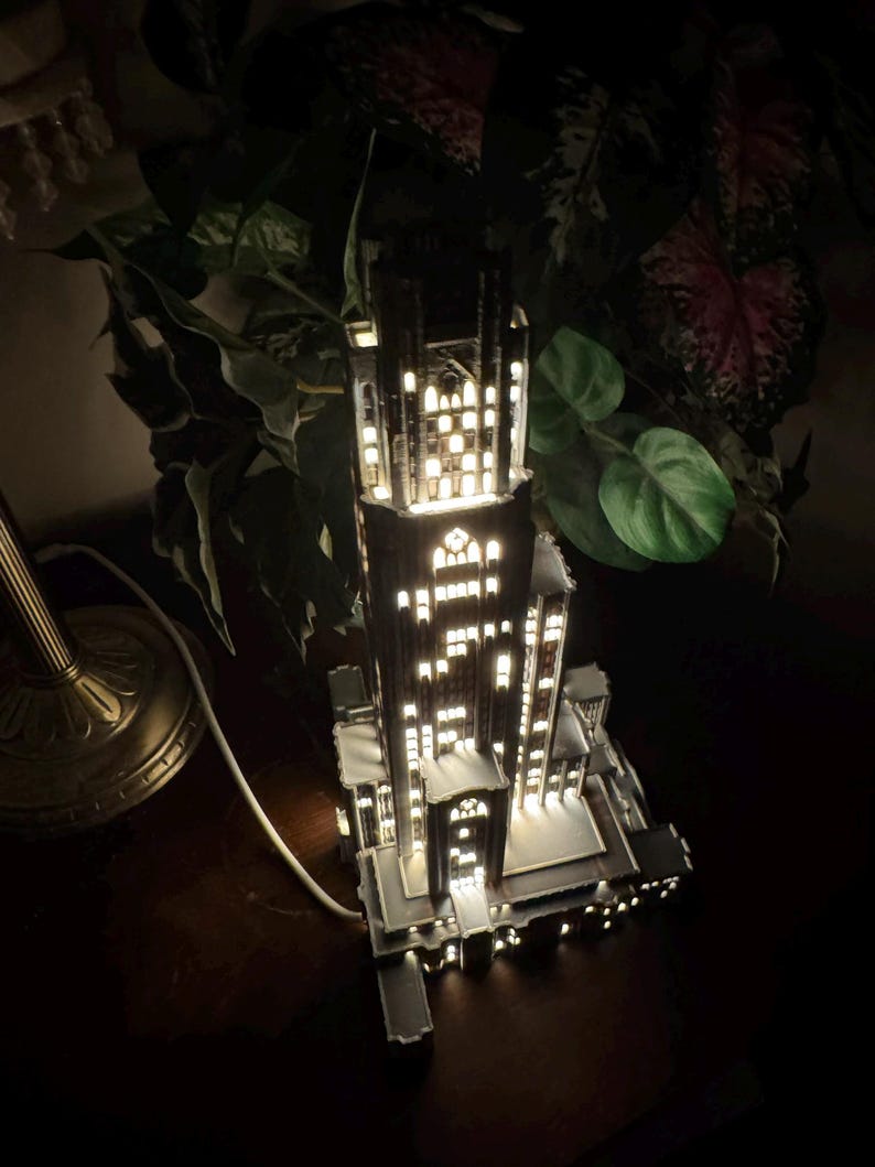 May include: A tall, dark, multi-tiered architectural lamp shaped like a skyscraper with numerous illuminated rectangular windows. The structure features intricate vertical detailing, a central tower, and a white power cord extending from the base.