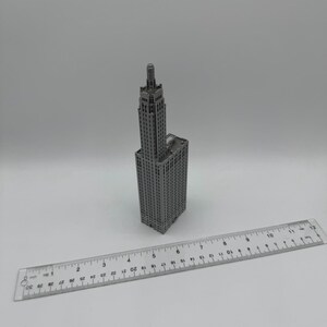 May include: A grey miniature model of a tall skyscraper with a pointed roof. The model is approximately 12.5 cm tall and is sitting on a white surface next to a ruler.