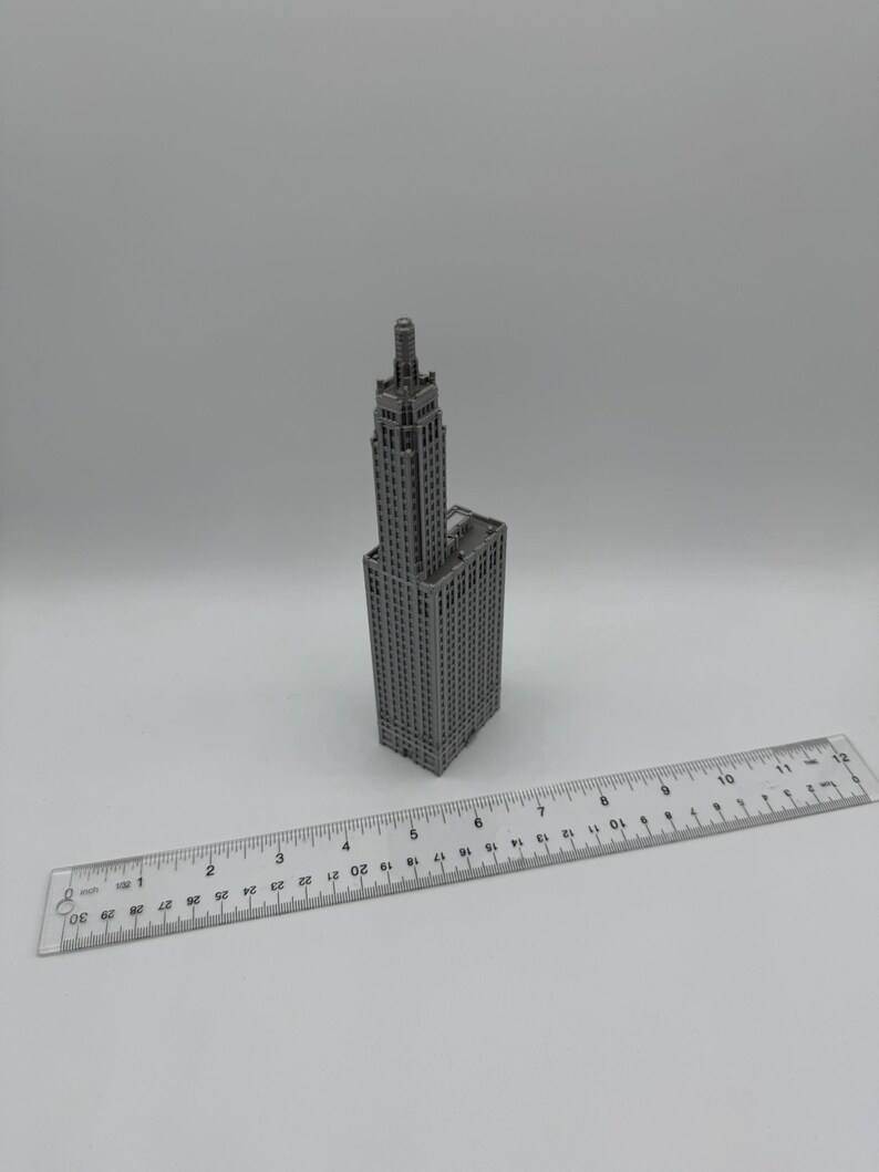 May include: A grey miniature model of a tall skyscraper with a pointed roof. The model is approximately 12.5 cm tall and is sitting on a white surface next to a ruler.