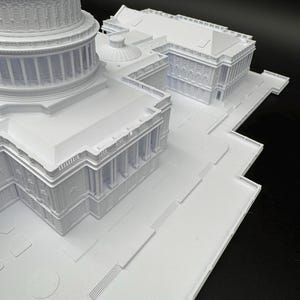 K&ouml;nnte beinhalten: Ein wei&szlig;es 3D-gedrucktes Modell eines Geb&auml;udekomplexes mit detaillierten architektonischen Elementen wie S&auml;ulen, Kuppeln und Treppen. Das Modell ist vor einem dunklen Hintergrund platziert, der das komplizierte Design hervorhebt.