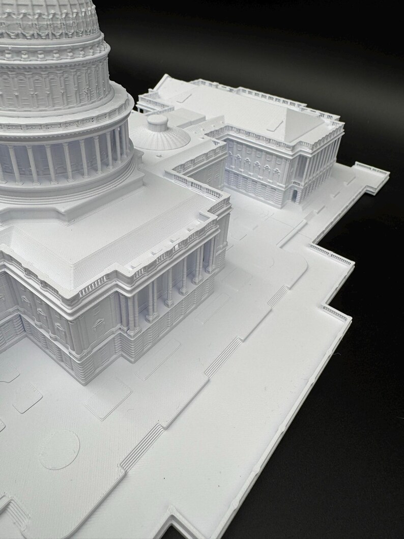 K&ouml;nnte beinhalten: Ein wei&szlig;es 3D-gedrucktes Modell eines Geb&auml;udekomplexes mit detaillierten architektonischen Elementen wie S&auml;ulen, Kuppeln und Treppen. Das Modell ist vor einem dunklen Hintergrund platziert, der das komplizierte Design hervorhebt.