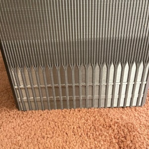 Pu&ograve; includere: Primo piano di un oggetto rettangolare grigio strutturato con linee verticali. L'oggetto ha una serie di linee parallele che convergono nella parte inferiore. L'oggetto &egrave; su una superficie marrone.