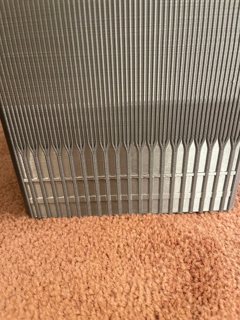 Pu&ograve; includere: Primo piano di un oggetto rettangolare grigio strutturato con linee verticali. L'oggetto ha una serie di linee parallele che convergono nella parte inferiore. L'oggetto &egrave; su una superficie marrone.