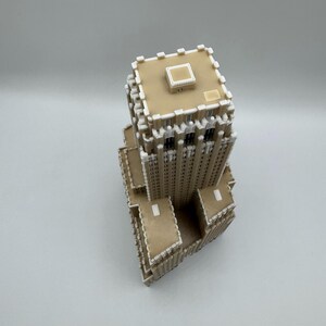 May include: A tan and white architectural model of a skyscraper. The model has a rectangular base and a detailed facade with multiple windows. The top of the building features a flat roof with a small square structure. The model is made of interlocking pieces.