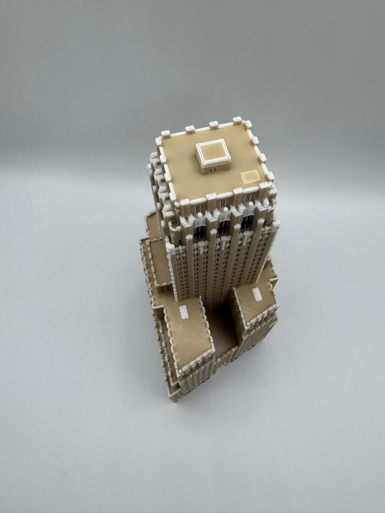 May include: A tan and white architectural model of a skyscraper. The model has a rectangular base and a detailed facade with multiple windows. The top of the building features a flat roof with a small square structure. The model is made of interlocking pieces.
