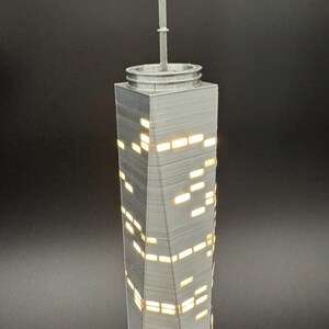 May include: A gray, 3D-printed model of a skyscraper with illuminated windows. The building has a spire at the top and a textured surface. The windows are rectangular and emit a warm, yellow light, creating a contrast against the gray structure.