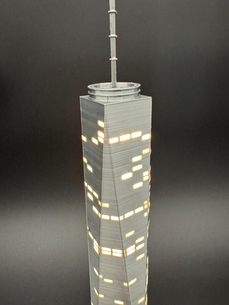 May include: A gray, 3D-printed model of a skyscraper with illuminated windows. The building has a spire at the top and a textured surface. The windows are rectangular and emit a warm, yellow light, creating a contrast against the gray structure.