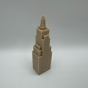 May include: A beige, miniature skyscraper model with a detailed, multi-tiered design. The building has a rectangular base and tapers upwards, featuring numerous windows and architectural details. The model stands approximately 8 inches tall.