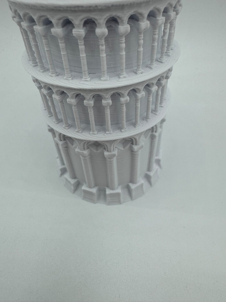 May include: A white 3D printed model of the Leaning Tower of Pisa. The model is detailed with arches and columns.