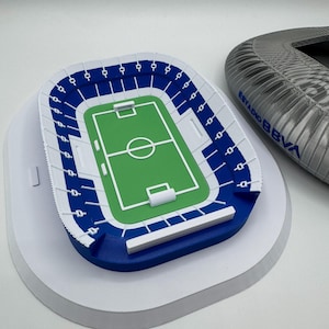 May include: A miniature stadium model with a green field, blue seating, and white accents. The model is on a white base. A partial stadium structure with the text "ESTADIO BBVA" is visible in the background.