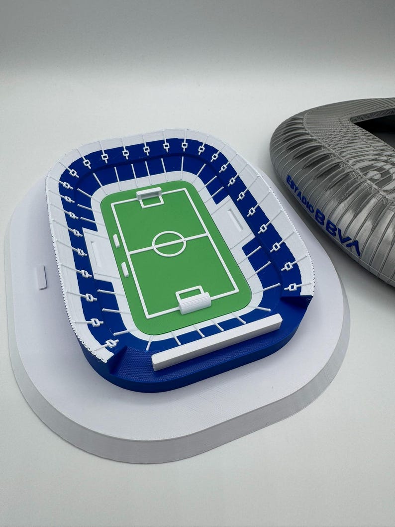 May include: A miniature stadium model with a green field, blue seating, and white accents. The model is on a white base. A partial stadium structure with the text "ESTADIO BBVA" is visible in the background.