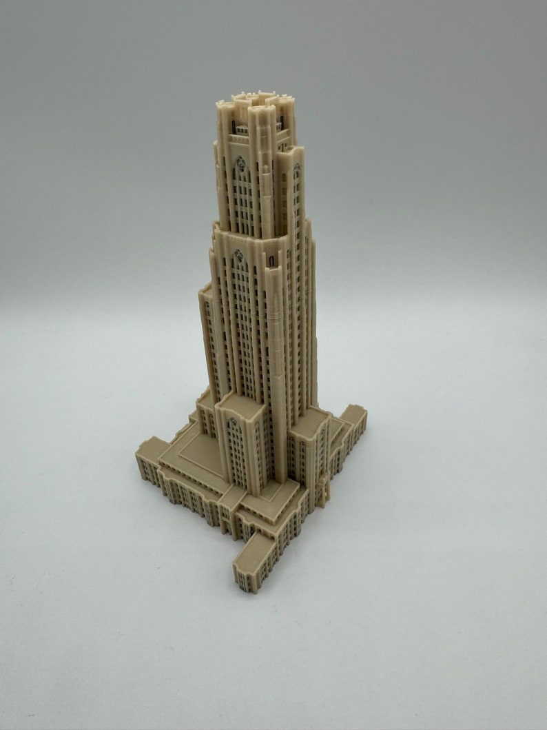 May include: A detailed 3D printed model of a tall, beige building with a pointed roof and many windows. The building has a rectangular base and is set against a plain white background.
