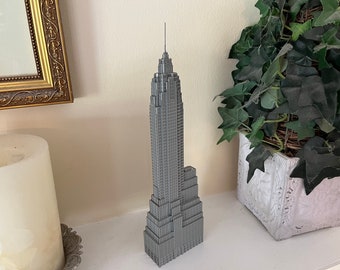 May include: A gray miniature replica of the Empire State Building. The model has intricate details, including windows and architectural features. The building stands tall on a white surface, with a green plant in a white pot to the right.