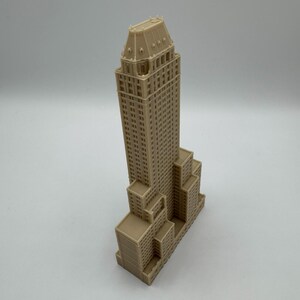 Może przedstawiać: Beżowy miniaturowy model wieżowca z szczeg&oacute;łowymi elementami architektonicznymi. Model ma wysoką, prostokątną konstrukcję gł&oacute;wną z wieloma oknami i szczeg&oacute;łowym dachem. Podstawa budynku ma kilka mniejszych prostokątnych sekcji.
