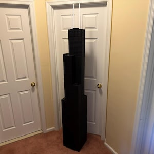 May include: A black architectural model of a skyscraper, with multiple rectangular sections stacked vertically. The model is positioned between two white doors and against a light yellow wall. The model is approximately 152 cm tall.