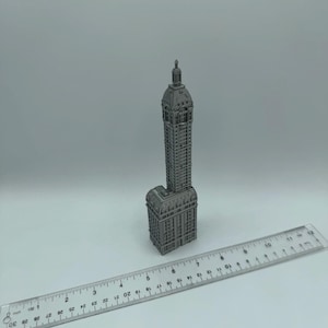 May include: A miniature model of a tall, gray building with a pointed roof and many windows. The model is made of metal and is sitting on a white surface next to a ruler.